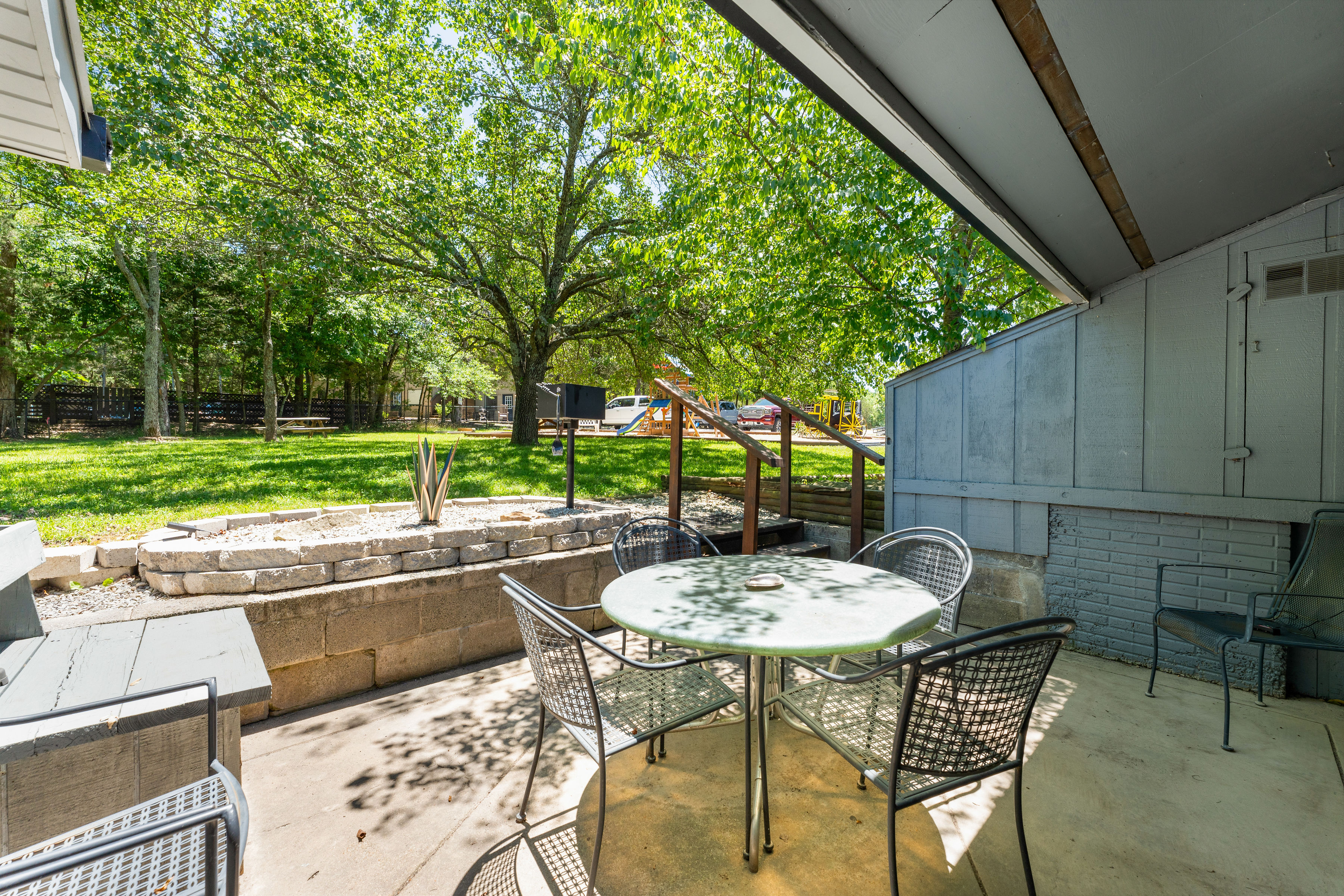A patio area with a metal table and chairs, a raised garden bed, and trees in the background.