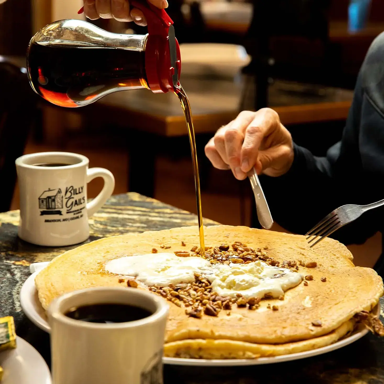 A hand pouring syrup on a pancake with butter and nuts at a table with a coffee cup.